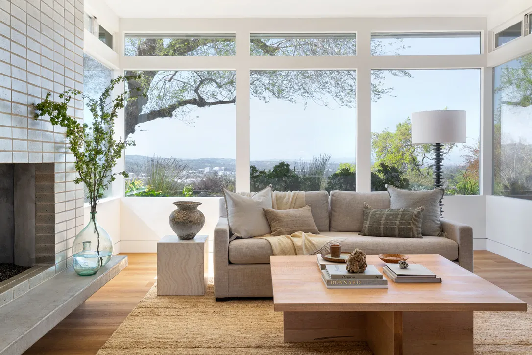 A modern living room featuring a beige sofa, a wooden table with books, a vase with greenery, large windows with scenic views, and a minimalist design, in a home by Gar-Cal Corporation in Livermore.